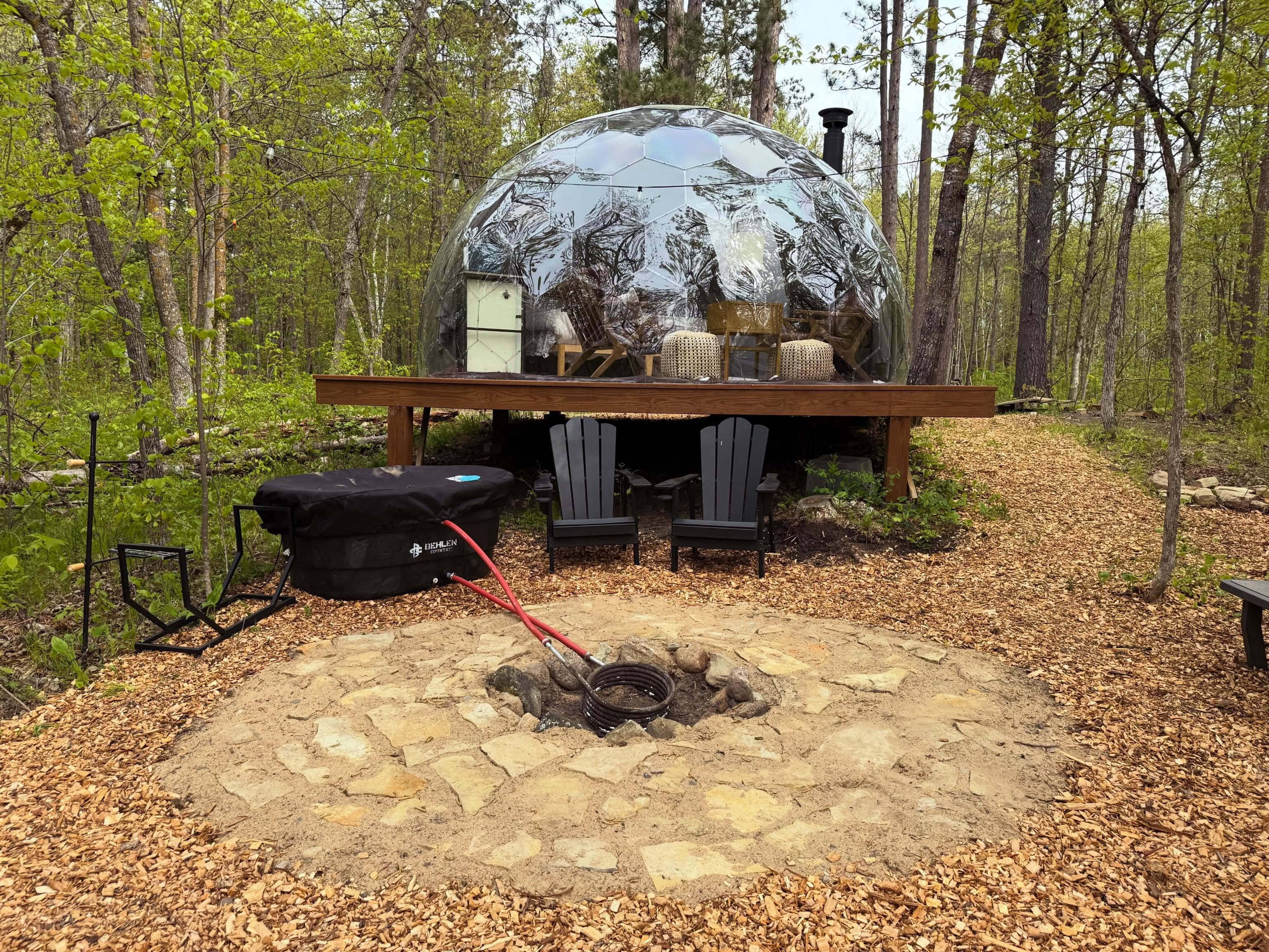Wide-angle view of geodesic dome in forest clearing with fire pit